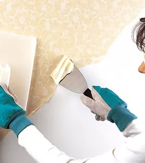 A person with gloved hands removes wallpaper using a scraper