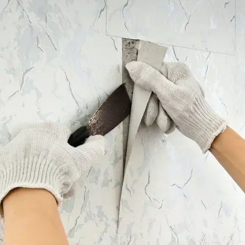 Two hands wearing gray gloves are using a putty knife to peel wallpaper