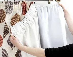 person hangs a white curtain next to a patterned curtain with leaf designs